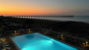 A swimming pool with lounge chairs and a view of a pier extending into the ocean at sunset.