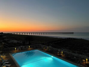 Una piscina con sillones de descanso y vistas a un muelle que se adentra en el océano al atardecer.