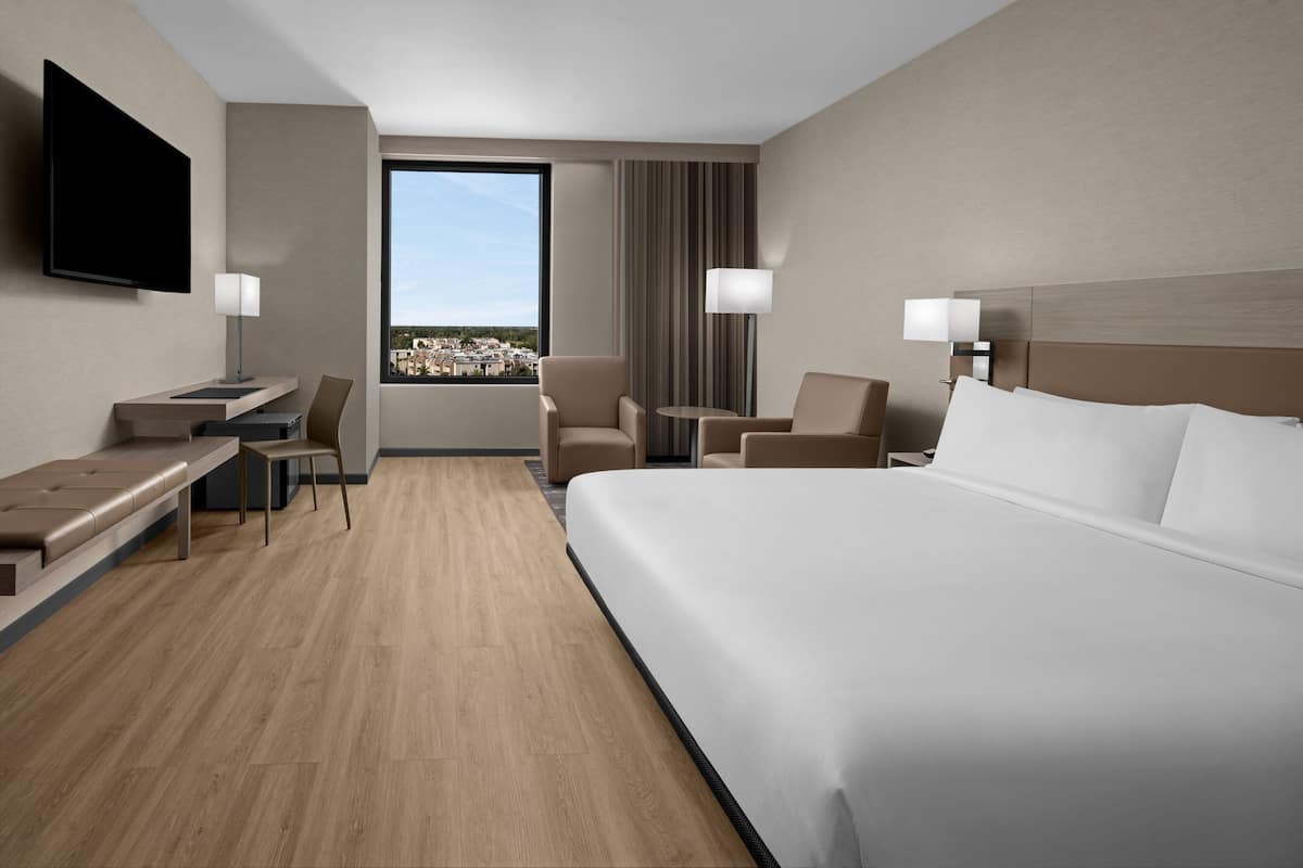 Ein Hotelzimmer mit einem großen Bett, einem Schreibtisch mit Stuhl, einem an der Wand befestigten Fernseher und einem Fenster mit Blick auf Gebäude.