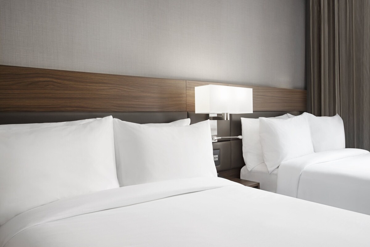 Une chambre d’hôtel avec une tête de lit en bois, deux lits avec du linge de lit blanc, une table de chevet et une lampe.