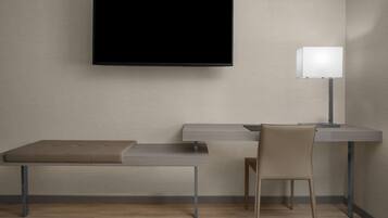 A modern room with a flat-screen TV mounted on the wall, a desk with a chair, and a bedside table with a lamp.
