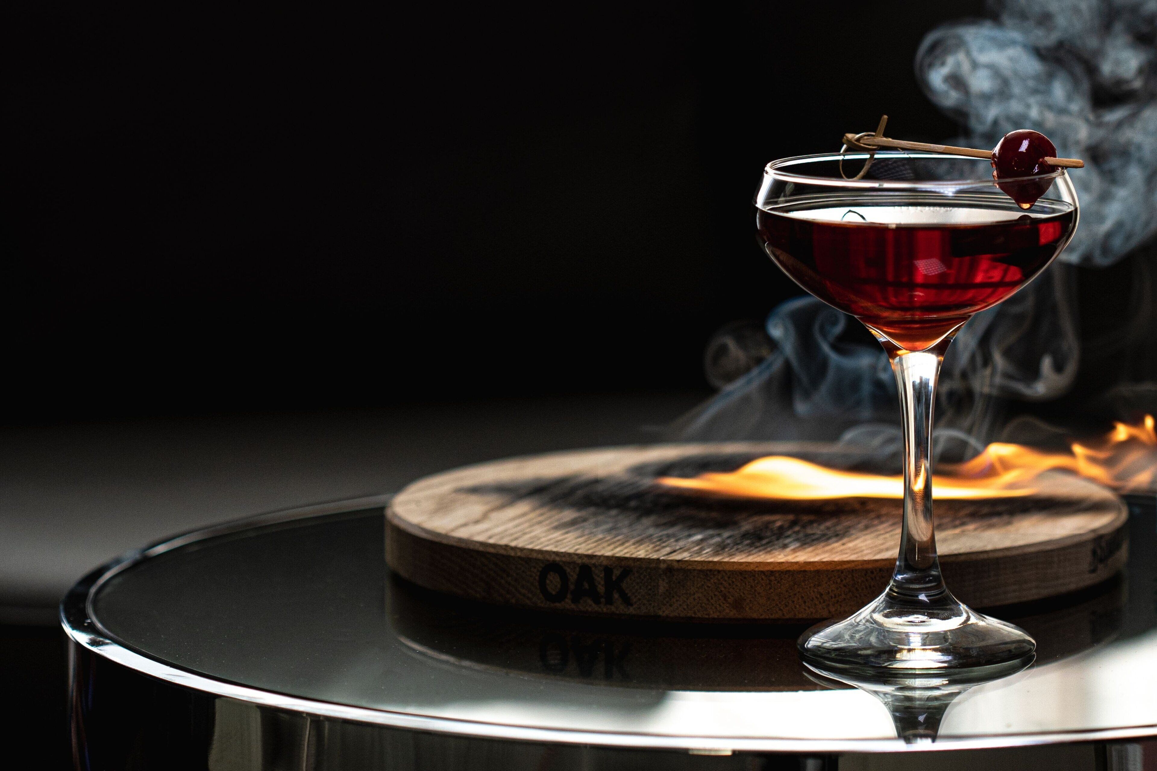 Martini-lasi, jossa on kirsikka varrastikussa, on asetettu pyöreän pöydän päälle. Pöydällä on puinen alusta, jossa lukee teksti ”OAK”.