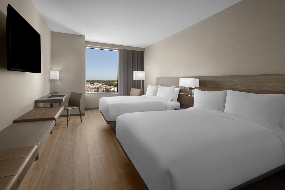 Ein Hotelzimmer mit zwei Betten, einem Schreibtisch, einem Stuhl, einem Fernseher und einem großen Fenster mit Blick auf die Stadt.