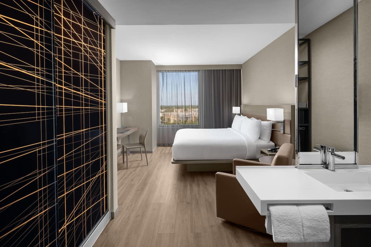 Ein modernes Hotelzimmer mit einem großen Bett, einem Schreibtisch, einem Stuhl, einem Badezimmer mit Waschbecken und Spiegel sowie einem Blick auf die Stadt durch das Fenster.