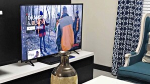A living room with a television displaying a show, a decorative vase on a table, and a patterned curtain in the background.