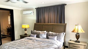 A bedroom with a bed, two bedside tables, a nightstand, a lamp, an air conditioning unit, and a ceiling fan.