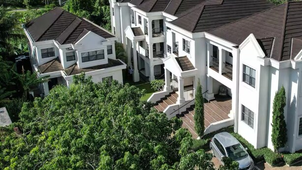 A large, white, multi-story residential building with a dark roof, surrounded by lush greenery and hills in the background.