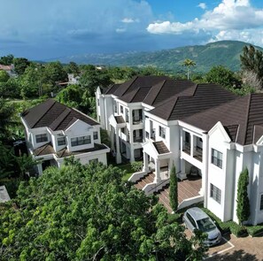 A large, white, multi-story residential building with a dark roof, surrounded by lush greenery and hills in the background. - Coconut Palms Luxury Apartment King Bed/ A/C,Gym and Pool (Manchester Parish)