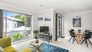 Uma sala de estar moderna com uma janela ampla, uma televisão de tela plana, uma área de jantar com mesa de vidro e um sofá.