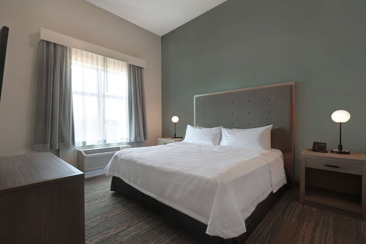 A hotel room with a large bed, two bedside tables with lamps, a window with curtains, and an air conditioning unit.