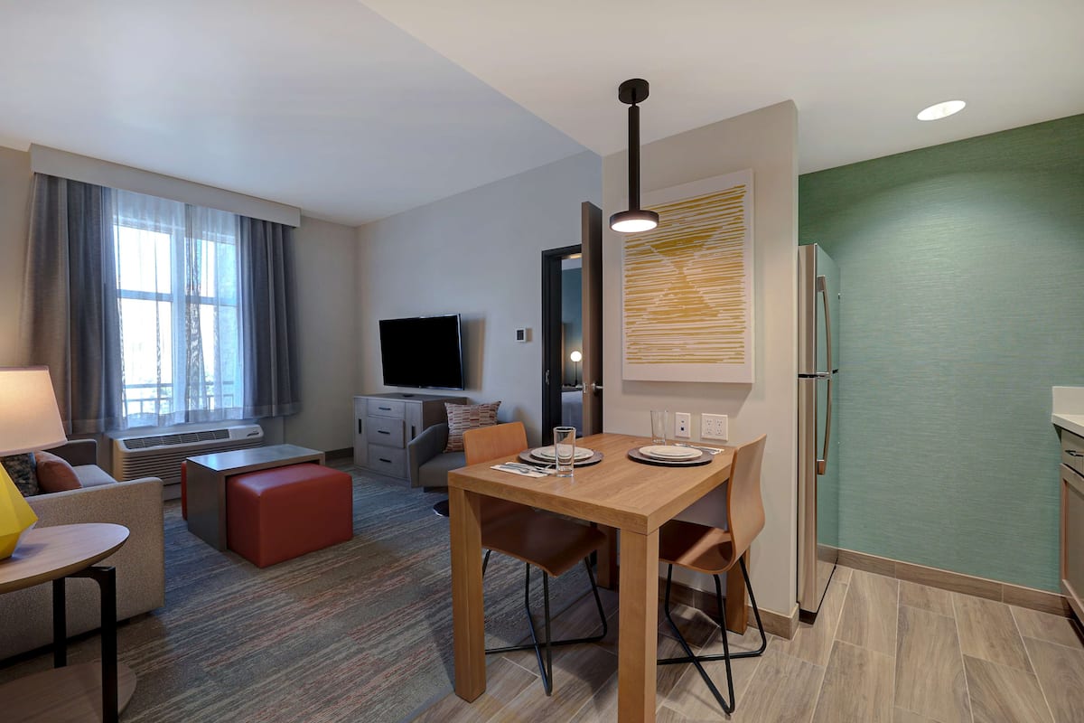 A modern hotel room with a dining area, a sofa, a TV, and a refrigerator.