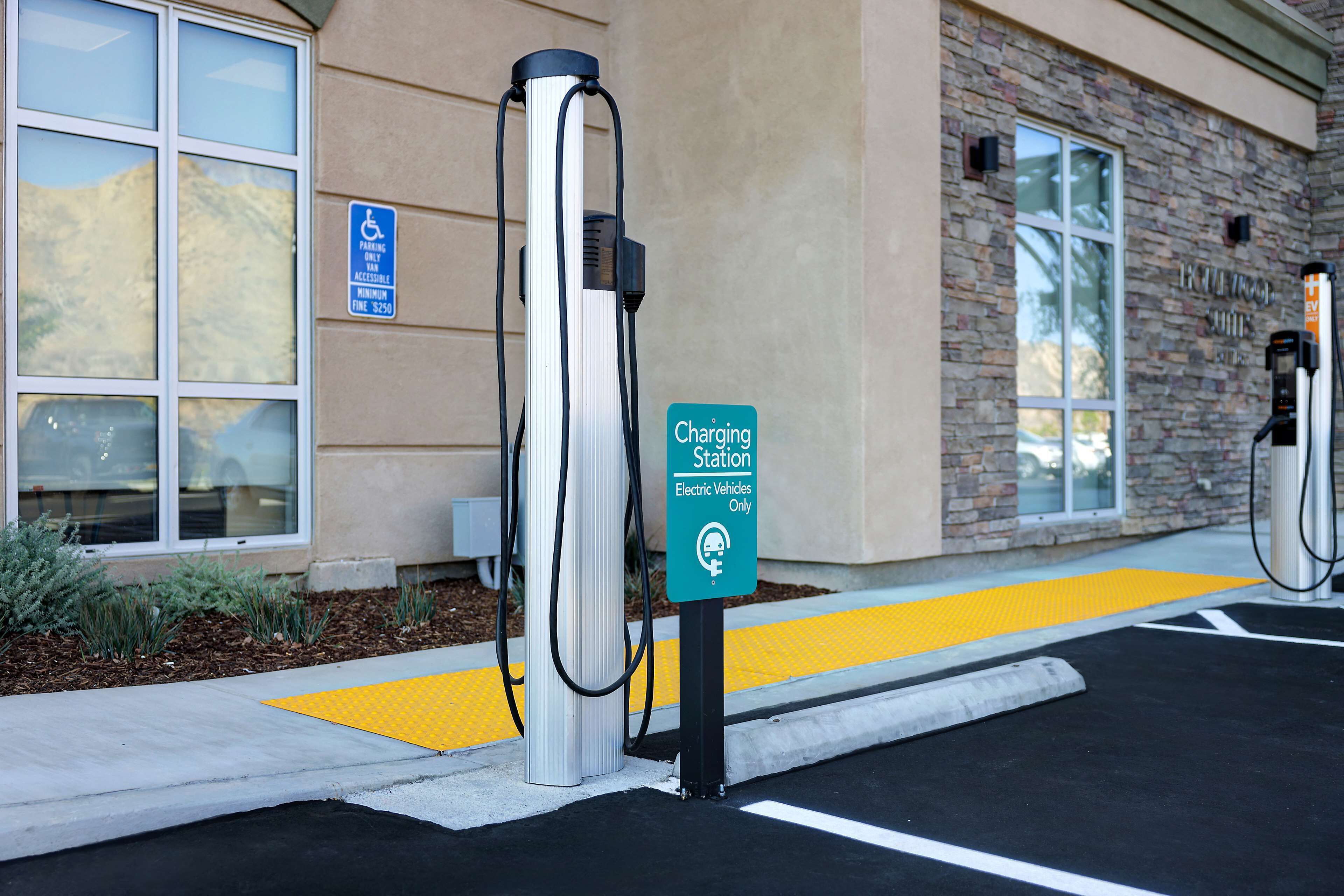 Une borne de recharge pour véhicules électriques, un panneau indiquant une place de stationnement réservée aux personnes handicapées et un bâtiment à la façade en pierre.