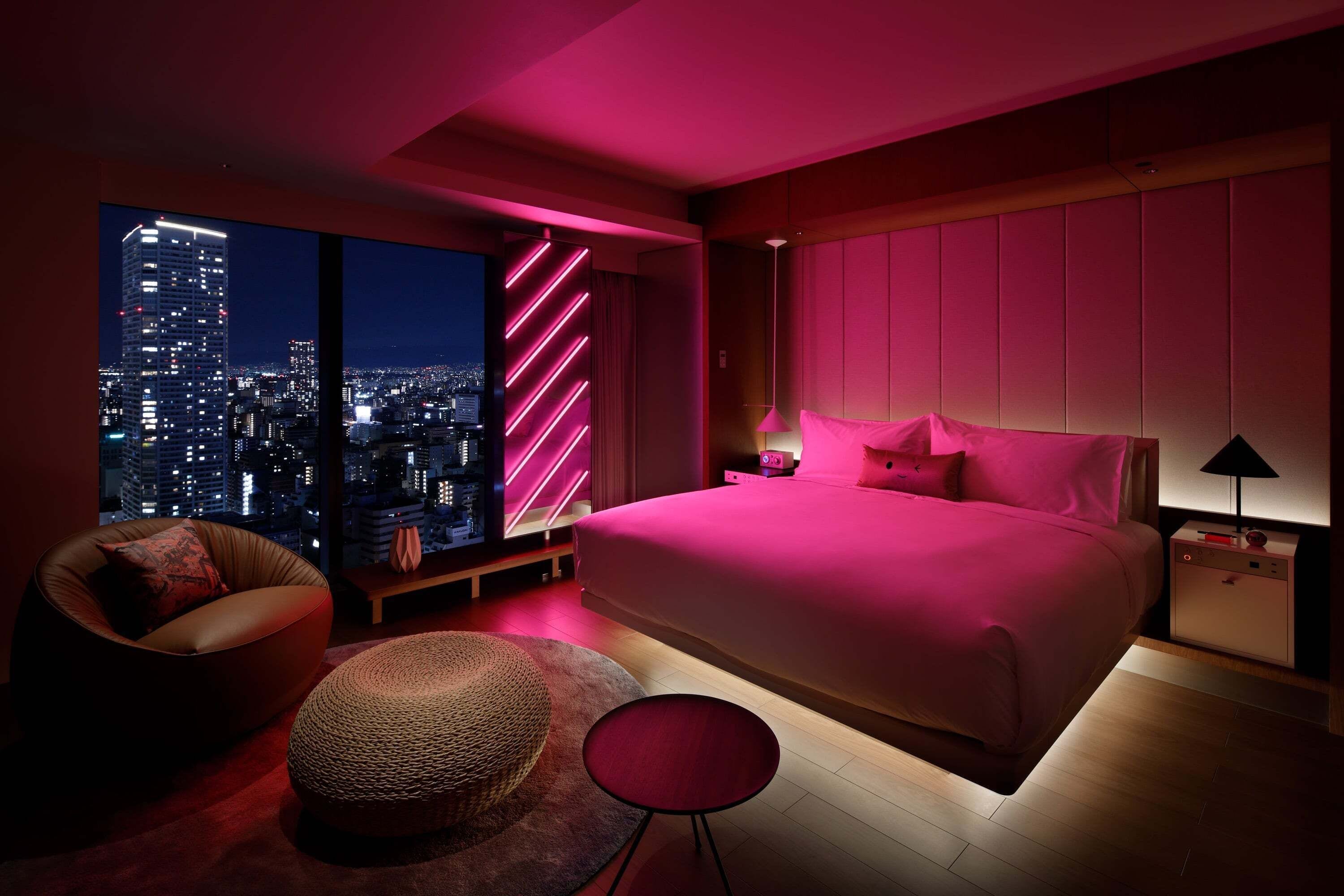 A modern bedroom with a large bed, a city view, and a pink-lit wall.