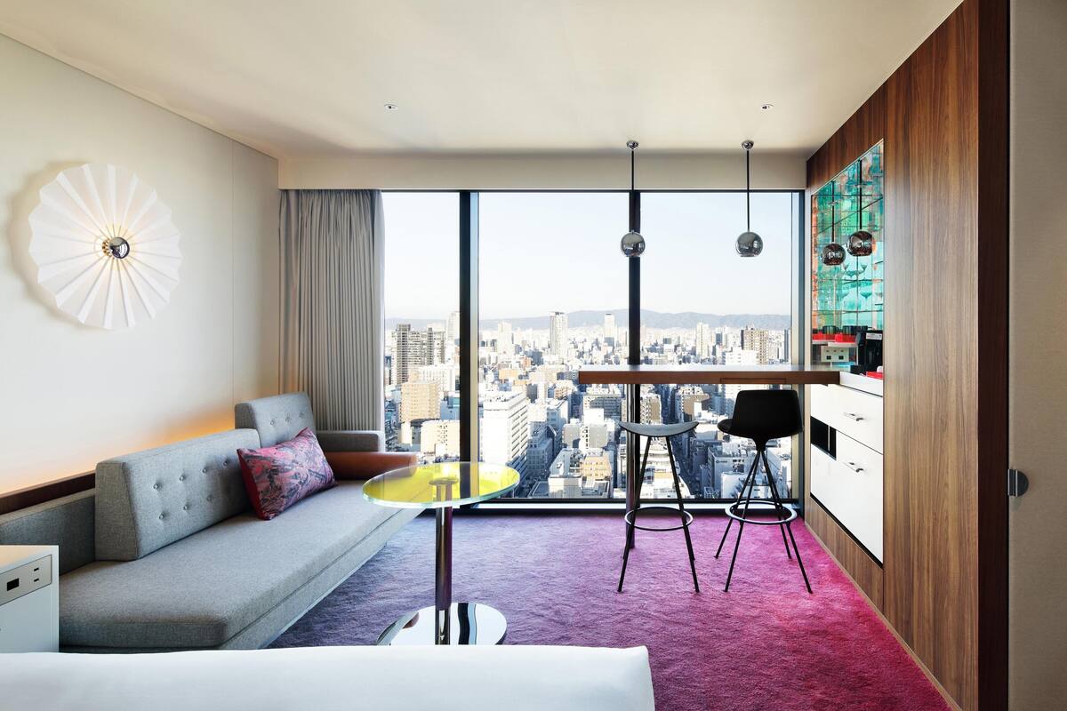 A modern hotel room with a city view, a grey sofa, a small yellow table, and a red carpet.