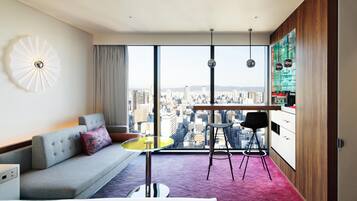 A modern hotel room with a city view, a grey sofa, a small yellow table, and a red carpet.