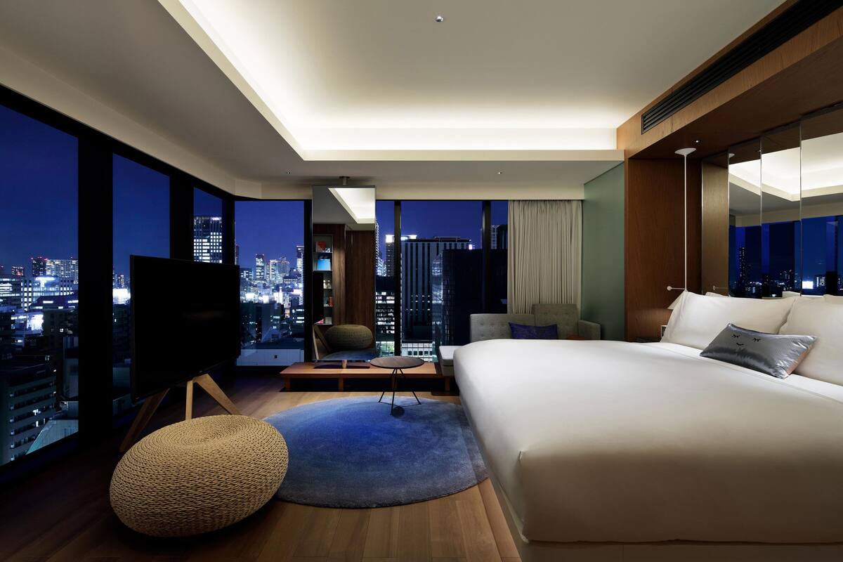 A modern hotel room with a large bed, a TV, a seating area, and a city view.