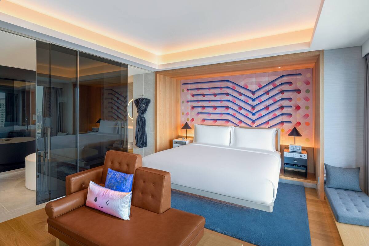A modern hotel room with a large bed, a glass-enclosed bathroom, a brown leather sofa, and a decorative wall panel.