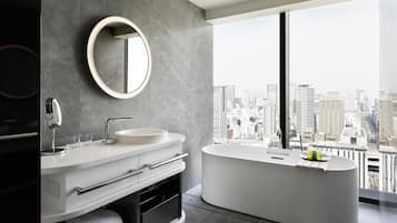 Un baño moderno con una bañera grande, dos lavamanos, un espejo redondo y vistas a la ciudad a través de ventanas amplias.