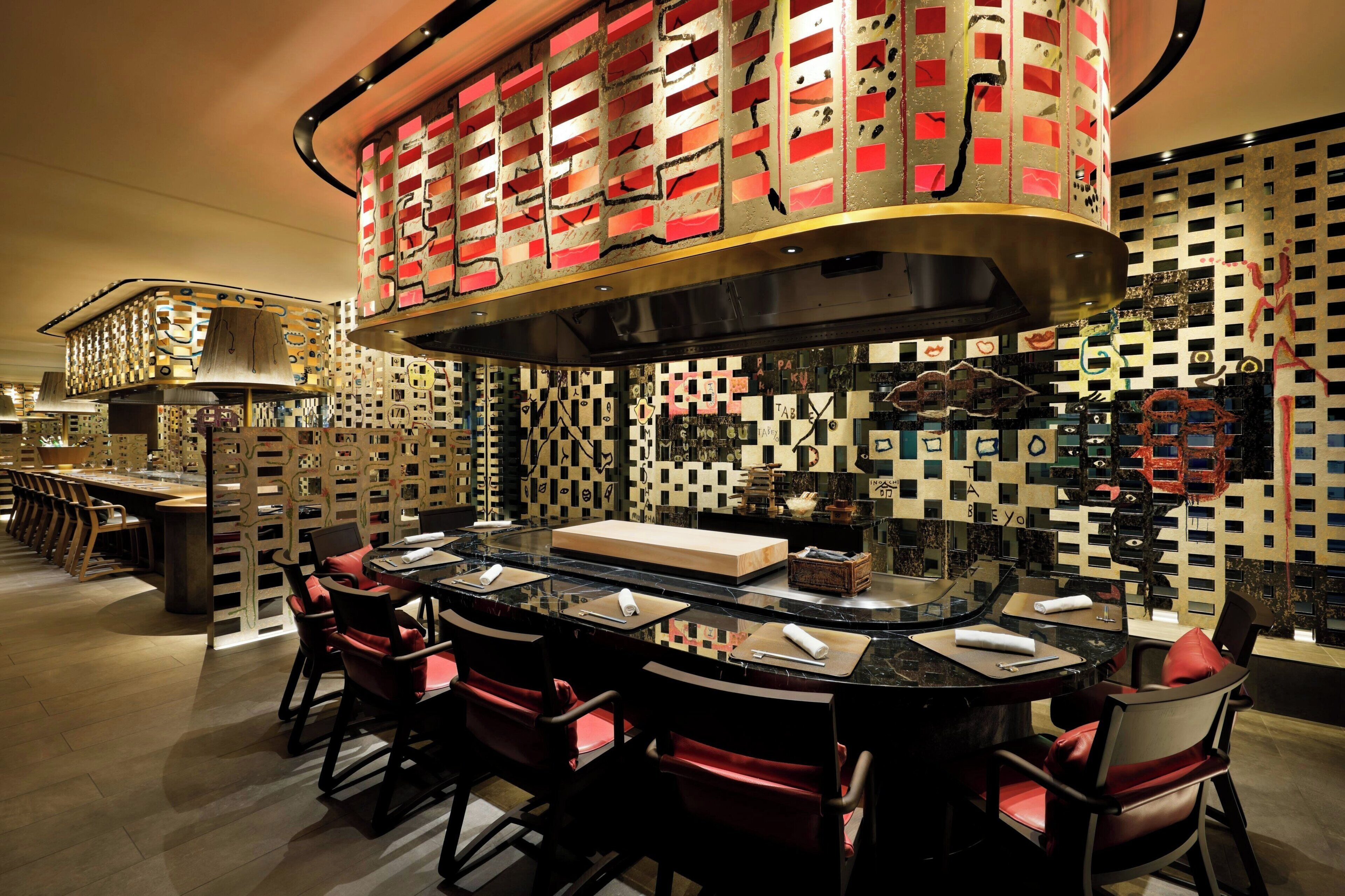 A modern restaurant with a central island, red upholstered chairs, and a patterned wall with various shapes and designs.