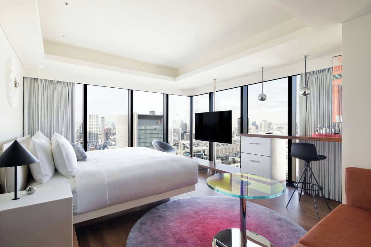 A modern hotel room with a large bed, a TV, a glass table, and a city view.