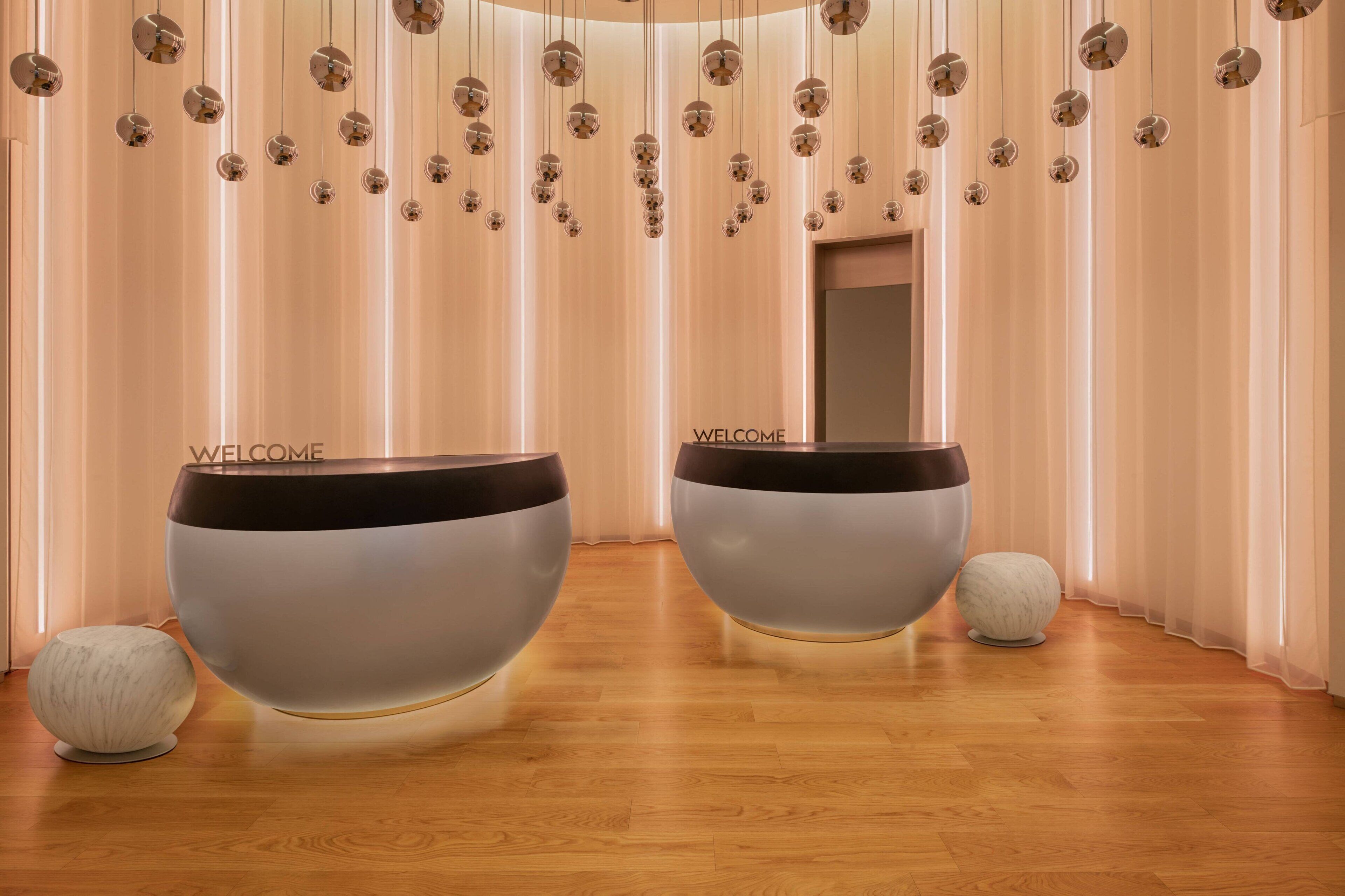 Two large, modern, bowl-shaped seating areas with 'WELCOME' text, a wooden floor, and a series of hanging spherical lights.