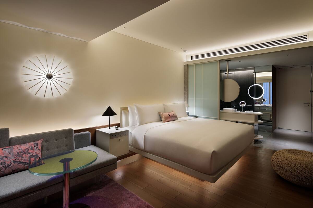 A modern bedroom with a large bed, a bedside table, a lamp, a round mirror, and a sofa with cushions.
