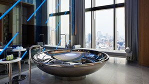 A modern bathroom with a large, round, metallic bathtub, a glass-enclosed shower area, and a city view through large windows. - W Osaka (Osaka)