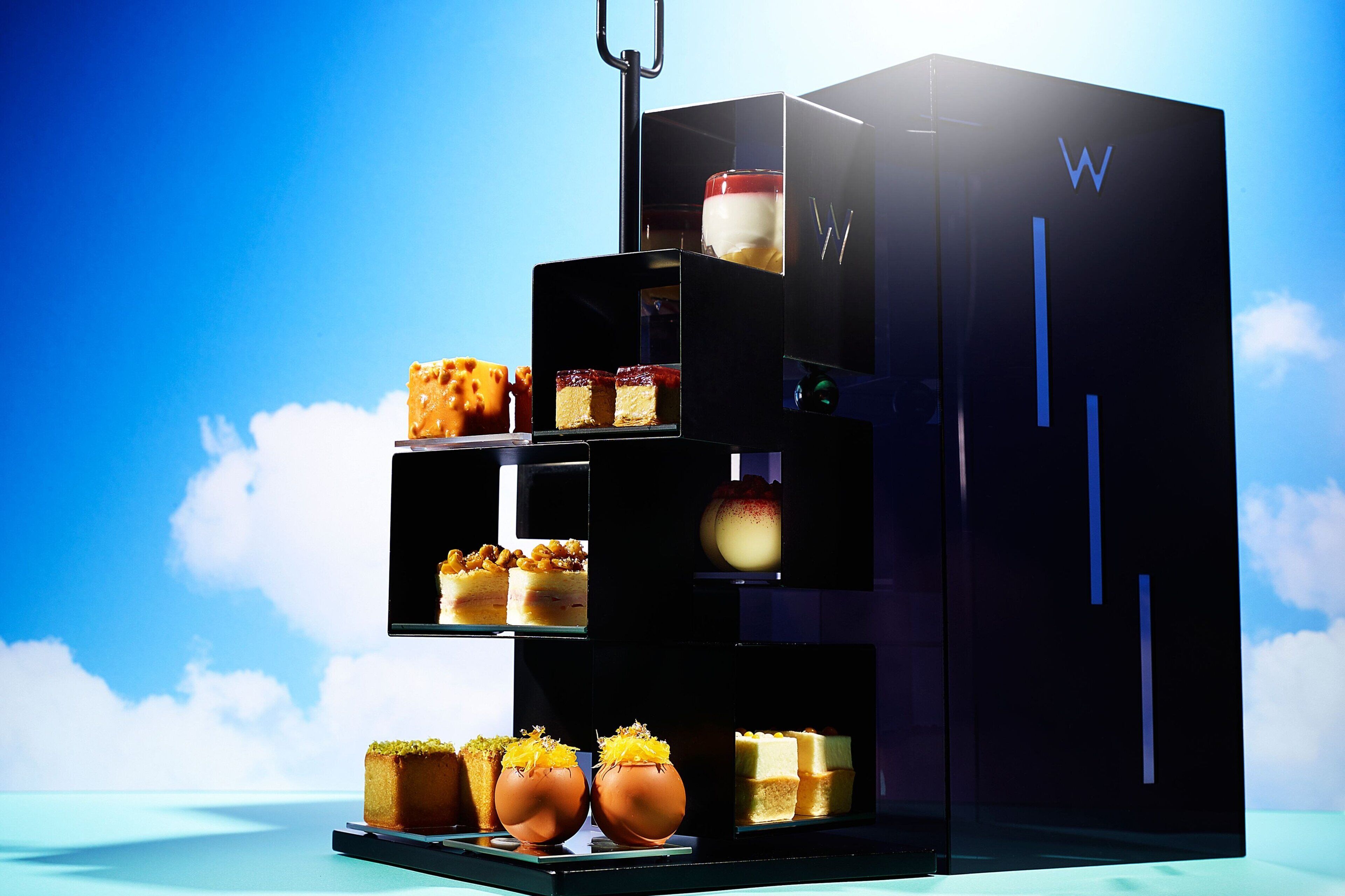 A modern, multi-tiered dessert display with various cakes and pastries, set against a clear blue sky.