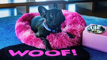 A black dog in a pink bed with a black mat on the floor that says "WOOF!".