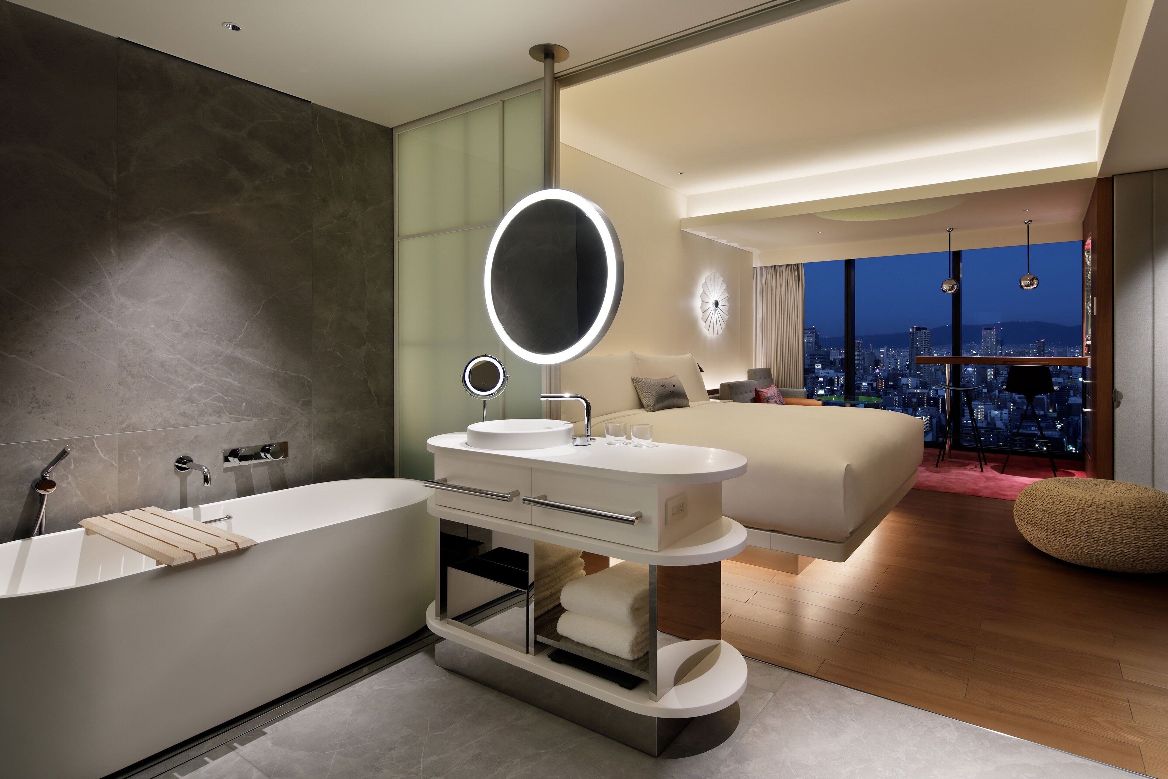 A modern bedroom with a large bed, a vanity with a mirror, and a view of a cityscape at night.