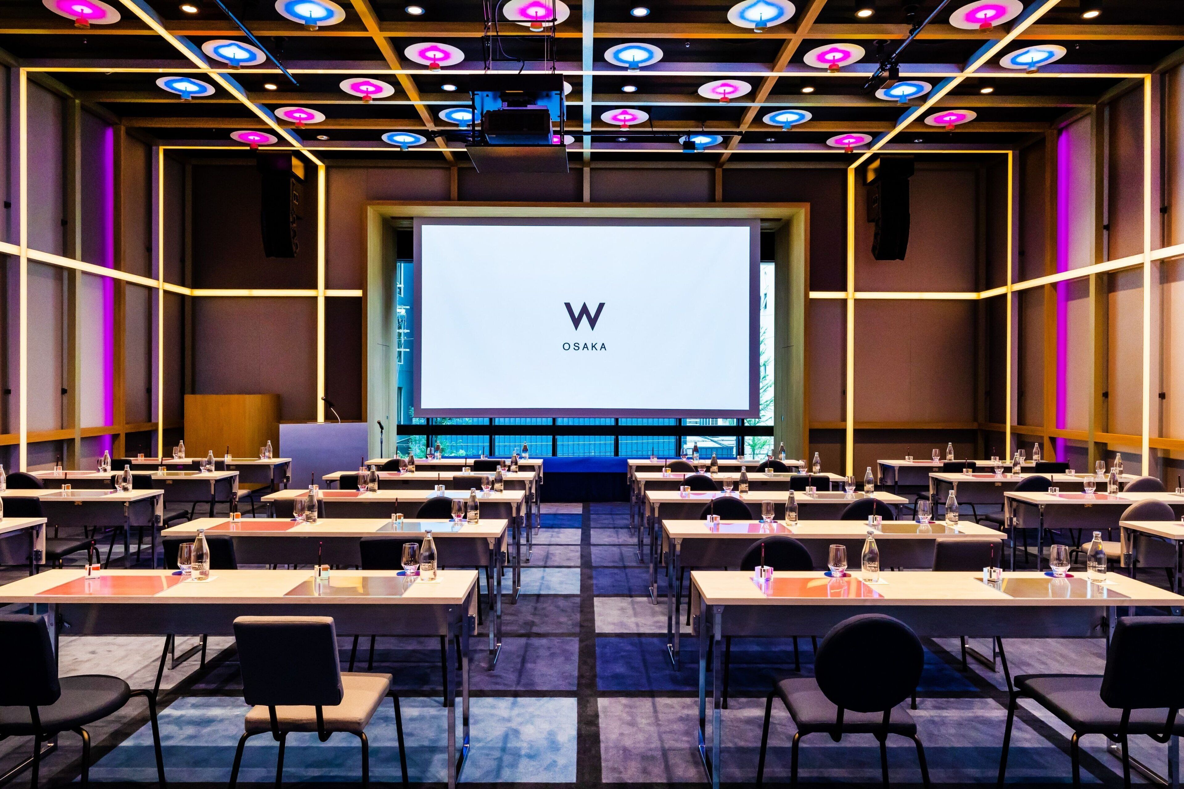 A conference room with a large screen displaying "W OSAKA" and multiple tables with chairs and water bottles.