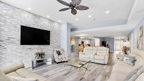 Un vaste salon doté d’un téléviseur à écran plat fixé sur un mur à texture, d’un ventilateur de plafond moderne et d’un coin repas situé à l’arrière-plan.