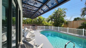 Une piscine entourée d’une clôture blanche, avec des fauteuils de jardin en osier et un espace abrité doté de panneaux de verre.
