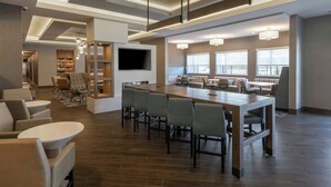 Un hall d’hôtel moderne avec une longue table commune, des coins salon et un téléviseur fixé au mur.