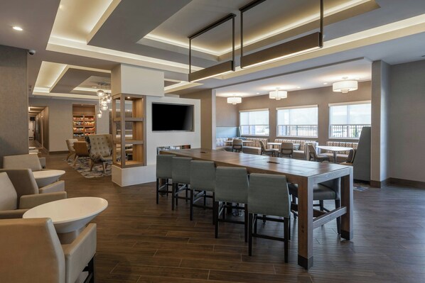A modern hotel lobby with a long communal table, seating areas, and a TV mounted on the wall.