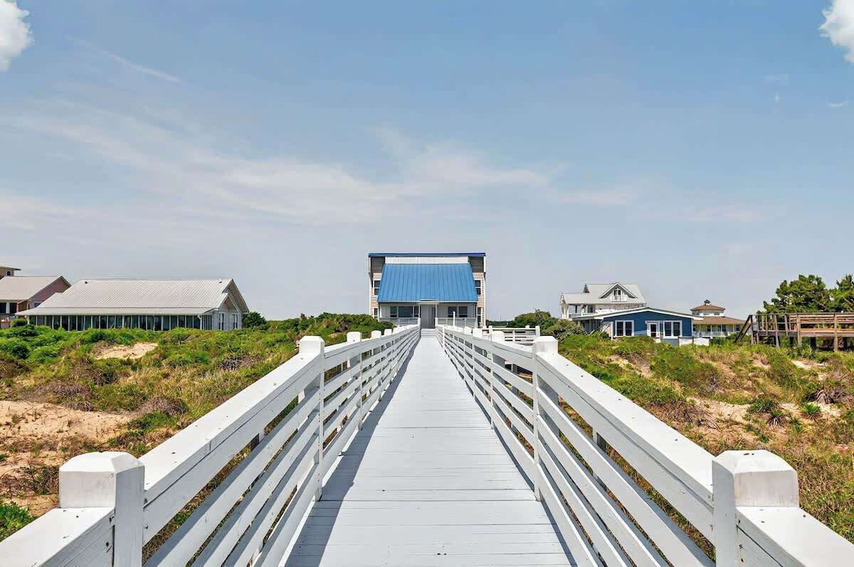 A wooden boardwalk leading to a building with a blue roof, surrounded by greenery and sandy terrain.