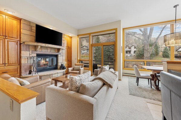 A modern living room with a fireplace, comfortable seating, a flat-screen TV, and large windows offering a view of the outdoors.