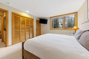 A bedroom with a large bed, wooden sliding doors, a TV mounted on the wall, and a window with a view of trees.