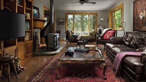 A cozy living room with a fireplace, leather sofa, wooden bookshelves, and a large window. - Stable View Cabin - Alpine Falls Ranch (Superior)