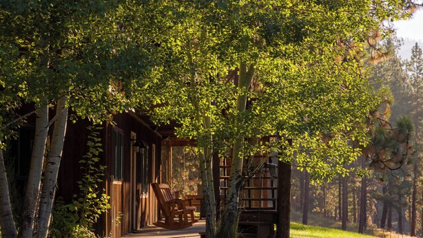 A wooden cabin with a porch, rocking chairs, and a bookshelf. - Stable View Cabin - Alpine Falls Ranch (Superior)