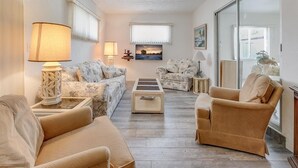 En stue med sofa, lænestole, et sofabord og lamper. Der er et vindue med udsigt, et billede på væggen og et spejl, der reflekterer rummet.