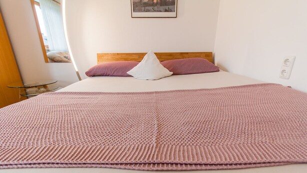 Een netjes opgemaakt bed met een wit-roze gestreept kussen, een houten hoofdbord en een grote roze gebreide deken.