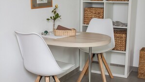 Een eetgedeelte met een ronde tafel, twee witte stoelen met houten poten, een rieten mand en een kleine potplant.
