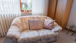Een woonkamer met een gestreept bankje, een kleine glazen tafel en uitzicht op het buitenland door een raam met dunne gordijnen.