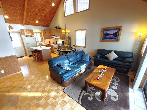 A spacious living area with a wooden ceiling, a dining table with chairs, a blue leather sofa, a dark leather sofa, and a kitchen in the background.