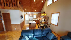A spacious living area with a black leather sofa, wooden ceiling, and a kitchen in the background.
