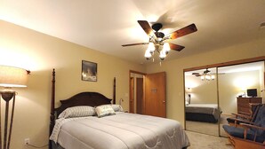 A bedroom with a bed, a ceiling fan, a nightstand, a lamp, a chair, and a mirror.