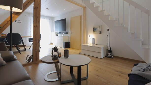 Una sala de estar moderna con una escalera, vigas de madera, una mesa redonda y un sofá.