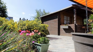 Una cabaña de madera con un porche, rodeada por un jardín con diversas flores y plantas.