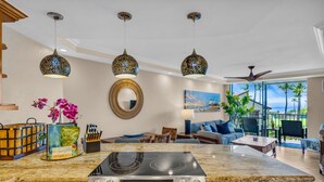 A modern kitchen with a central island, overhead pendant lights, and a large window offering a view of palm trees. - Elegant Oceanfront Vistas (Kihei)